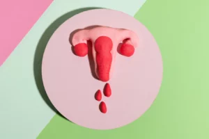 Ilustração de uma vagina em modelagem de argila com gotas vermelhas, representando o corrimento vaginal, em fundo de cores suaves. Melhor para entender sintomas e saúde íntima feminina.