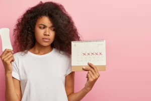 Jovem com expressão preocupada segurando um calendário com marcas de cruzes vermelhas e uma embalagem de absorvente, ilustrando irregularidade na menstruação.