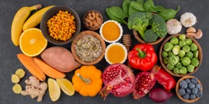 Variedade de alimentos ricos em potássio, incluindo bananas, brócolis, batata-doce, pimentão vermelho e frutas como romã e laranja, dispostos sobre uma superfície escura.