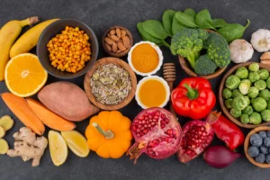 Variedade de alimentos ricos em potássio, incluindo bananas, brócolis, batata-doce, pimentão vermelho e frutas como romã e laranja, dispostos sobre uma superfície escura.