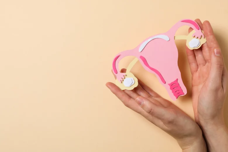 Ilustração de uma vagina de papel com detalhes em rosa, representando a saúde da mulher, sendo segurada por duas mãos femininas, sobre fundo bege.