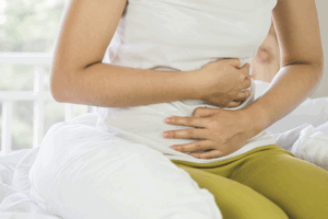 Mulher sentada na cama segurando a barriga com as duas mãos, representando os sintomas de cólica intestinal e diarreia.