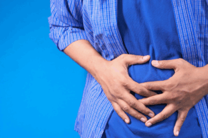 Homem com camisa azul segurando a barriga com as duas mãos, representando desconforto abdominal e problemas de saúde digestiva.